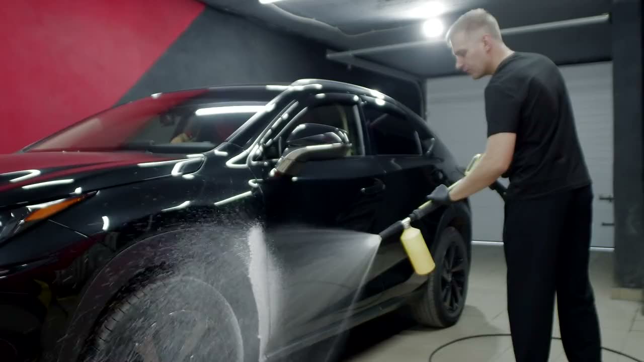 A glossy black SUV being covered in thick white wash foam inside a dark studio